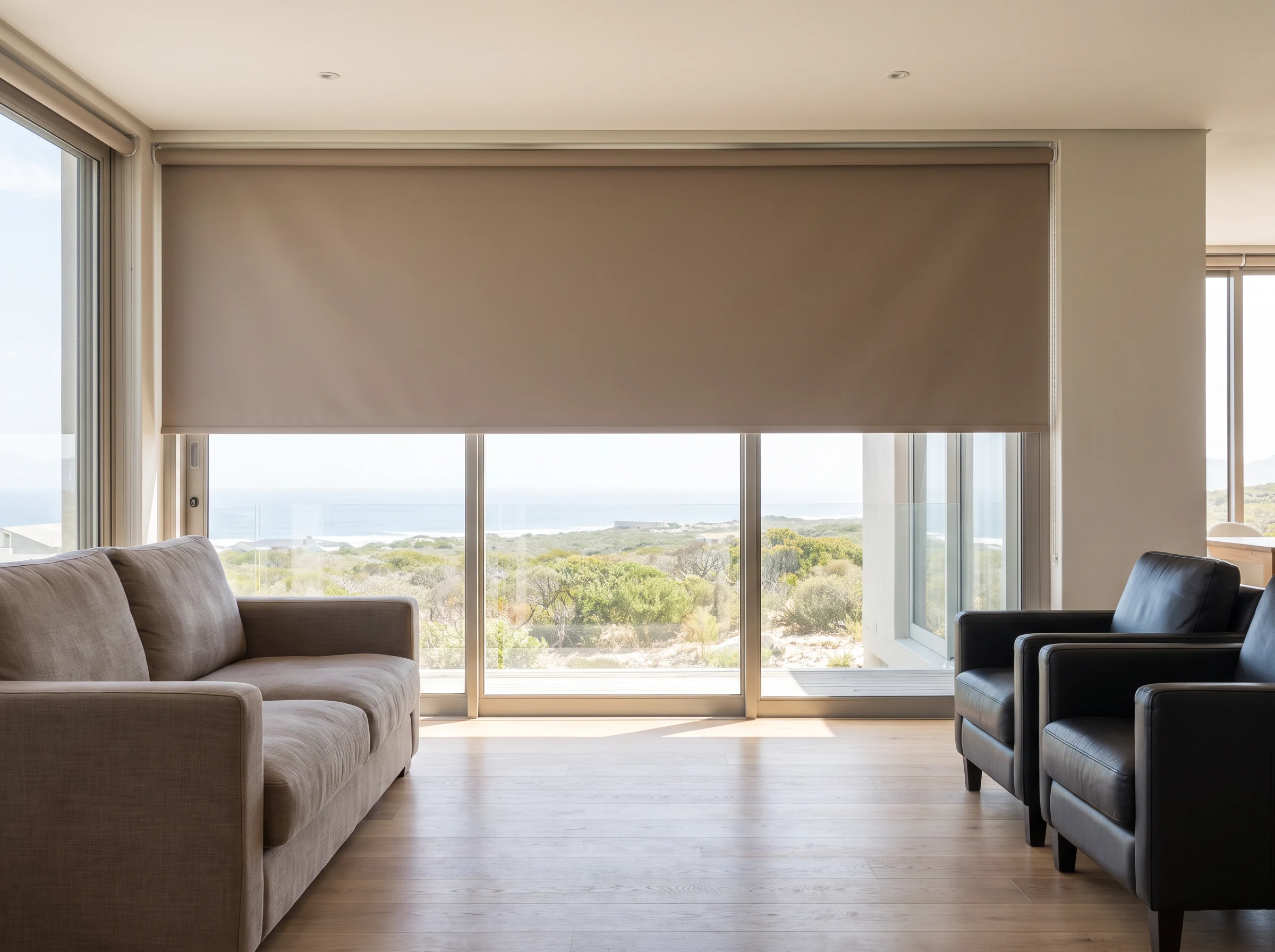 Taupe blockout roller blind lowered over a sliding door in a Sunningdale lounge with a coastal dune view beyond